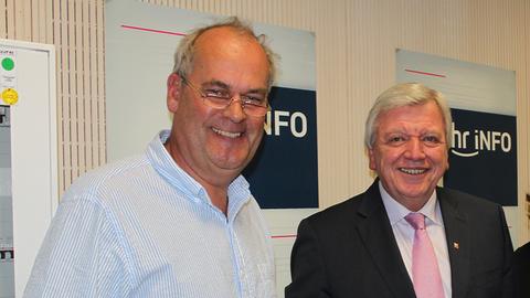 Christopher Plass und Volker Bouffier (Brustbild) im Studio vor hr-iNFO-Plakaten an der Wand, beide lachen in die Kamera, neueres Bild