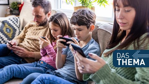 Mutter, Vater und zwei Kinder sitzen zusammen auf dem Sofa und alle schauen auf ihr Smartphone.