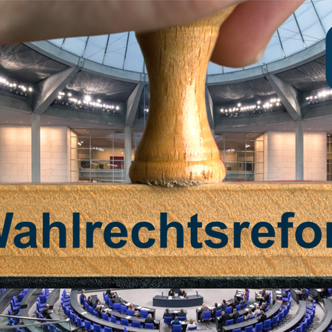 Ein symbolischer Holzstempel mit der Aufschrift "Wahlrechtsreform", gehalten von einer Hand, vor dem Plenum des Deutschen Bundestages im Reichstagsgebäude.