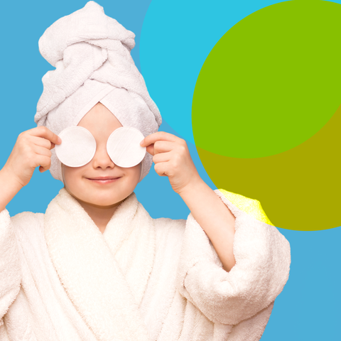 Ein Kind im Bademantel mit einem Handtuchturban auf dem Kopf, das sich zwei Wattepads vor die Augen hält und lächelt