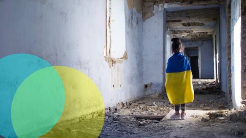 Ein Mädchen in einem zerstörten Haus, man sieht sie von hinten und über ihren Schultern hängt eine ukrainische Flagge