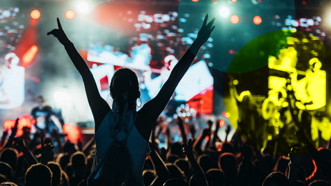 Eine Frau auf einem Konzert, die von hinten gezeigt wird. Sie sitzt auf den Schultern eines anderen Menschen und streckt beide Arme nach oben. Vor ihr ist eine Bühne