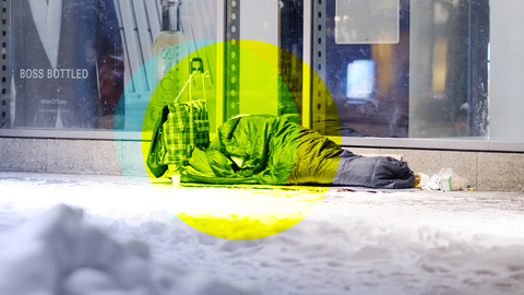 Ein obdachloser Mensch liegt bei Schnee im Schlafsack vor einem Schaufenster auf dem Gehweg. 