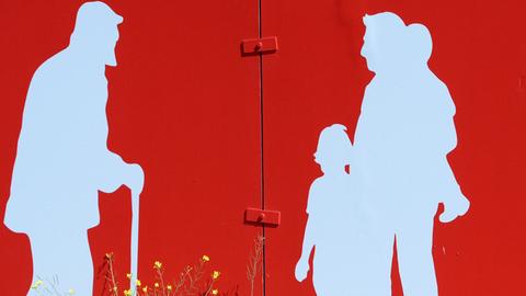 An einer roten Hauswand sieht man die weiße Silhouette eines Mannes am Gehstoch und ihm gegenüber ein Mann mit zwei Kindern