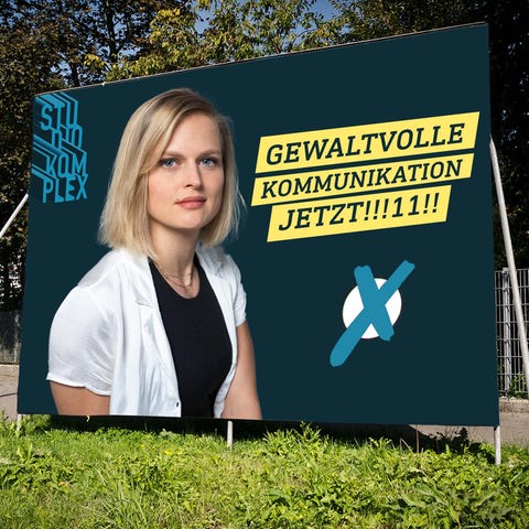 Auf dem Bild sieht man großes Wahlplakat auf der Straße auf dem die Hostin Anne den Betrachter vertrauenswürdig und offen anschaut. Als Wahlslogan steht: Gewaltvolle Kommunikation jetzt!!!11!!