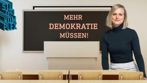 Die Hostin Anne blickt freundlich und gewinnend in ihre imaginäre Klasse, in der sich der Betrachter des Bildes selbst befindet. An der Tafel, die direkt und gut zu sehen ist, steht die Aufschrift: "Mehr Demokratie müssen!"