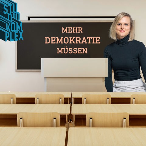 Die Hostin Anne blickt freundlich und gewinnend in ihre imaginäre Klasse, in der sich der Betrachter des Bildes selbst befindet. An der Tafel, die direkt und gut zu sehen ist, steht die Aufschrift: "Mehr Demokratie müssen!"