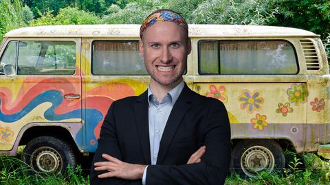 Der Host David trägt ein dunkelblaues klassisches Sakko und ein Hippie-Stirnband. Er steht mit verschränkten Armen und und einem breiten Grinsen vor einem Hippie-VW-Bus. 