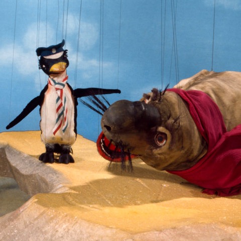 Das Bild zeigt einen Ausschnitt der Augsburger Puppenkiste. Links steht ein Pinguin mit einer blau-weiß-rot karrierten Krawatte und einer dunkel blauen Kapitänsmütze. Rechts liegt ein großes, grau-braunes Walross mit einem dunkelroten Schal. Der Hintergrund ist hellblau mit weißen Wolken und die beiden Puppen stehen auf einem beigen Steinvorsprung.