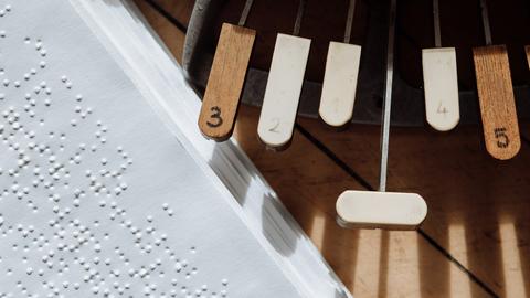 Draufsicht auf ein Buch mit Text in Braille, daneben eine einer Braille-Schreibmaschine auf einem braunen Tisch, der von der Sonne angestrahlt wird