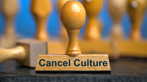 Ein Holzstempel mit der Aufschrift "Cancel Culture" auf einer Schieferplatte mit weiteren Stempeln unscharf dahinter vor einem blauen Hintergrund