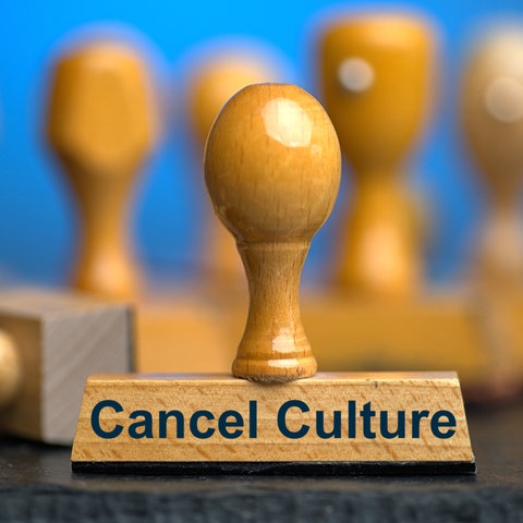 Ein Holzstempel mit der Aufschrift "Cancel Culture" auf einer Schieferplatte mit weiteren Stempeln unscharf dahinter vor einem blauen Hintergrund