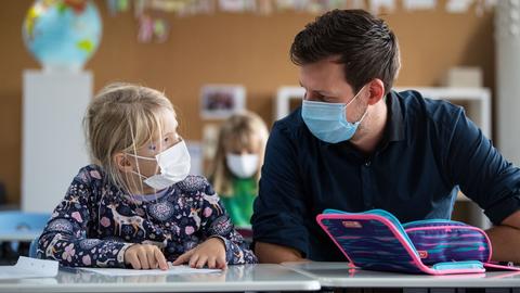 Schülerin und Lehrer sitzen mit Maske im Klassenraum nebeneinander