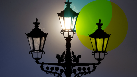 Eine schwarze Straßenlaterne mit drei Lampen leuchtet vor einem grauen Hintergrund. Vor zwei der Lampen wurden jeweils ein transparenter blauer und gelber Kreis gelegt.