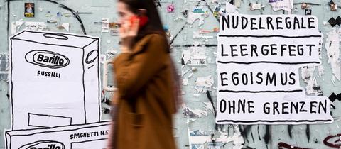 Eine Frau geht an einer Plakatwand vorbei, an der geschrieben steht "Nudelregale leergefegt - Egoismus ohne Grenzen". 