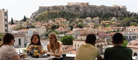 Menschen sitzen in einem Café, im Hintergrund die Akropolis