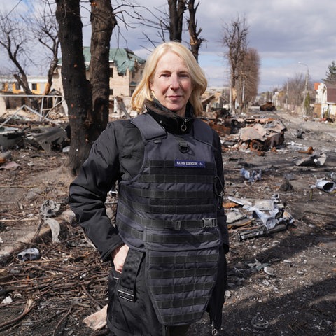 Frau in schwarzen Klamotten und mit blonden Haaren steht vor Trümmerfeld mit abgebrannten Bäumen. Sie trägt eine Schutzweste.