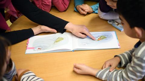 Kinder sitzen mit einer Erzieherin an einem Tisch und schauen ein Bilderbuch an