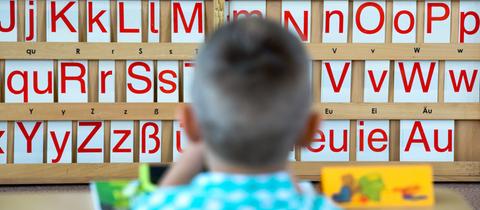 Ein Kind sitzt an einem Tisch, auf dem im Hintergrund Buchstaben zu sehen sind.