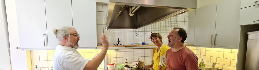 Industrieküche mit Kochinsel in der Mitte, Mann links würft Mann rechts etwas in den Mund, Frau rechts im Hintergrund schaut lachend zu