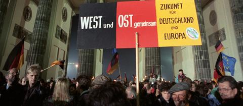 Feier Wiedervereinigung 1990 vor dem Brandenburger Tor: Feiernde halten scharz-rot-goldenes Transparent hoch: "West und Ost gemeinsam. Zukunft für Deutschland und Europa", im Hintergrund das Brandenburger Tor am Abend.