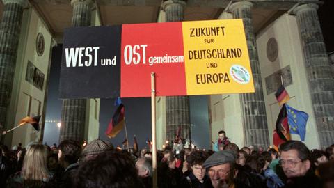 Feier Wiedervereinigung 1990 vor dem Brandenburger Tor: Feiernde halten scharz-rot-goldenes Transparent hoch: "West und Ost gemeinsam. Zukunft für Deutschland und Europa", im Hintergrund das Brandenburger Tor am Abend.