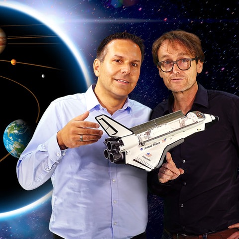 Mann mit hellblauem Hemd und kurzen braunen Haaren steht neben Mann mit mittellangen braunen Haaren, schwarzer Brille und schwarzem Hemd. Sie halten ein weißes Space Shuttle vor sich. Im Hintergrund ist in einem Kreis das Sonnensystem mit Planeten zu sehen.